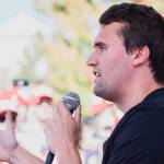 Charlie Kirk in a black T-shirt holding a microphone at an outdoor event.